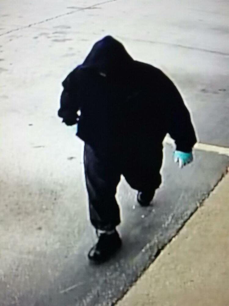 Camera footage of a man dressed in a black hoodie, black sweatpants, and black sneakers walking towards a building with his right hand reaching into his hoodie pocket.