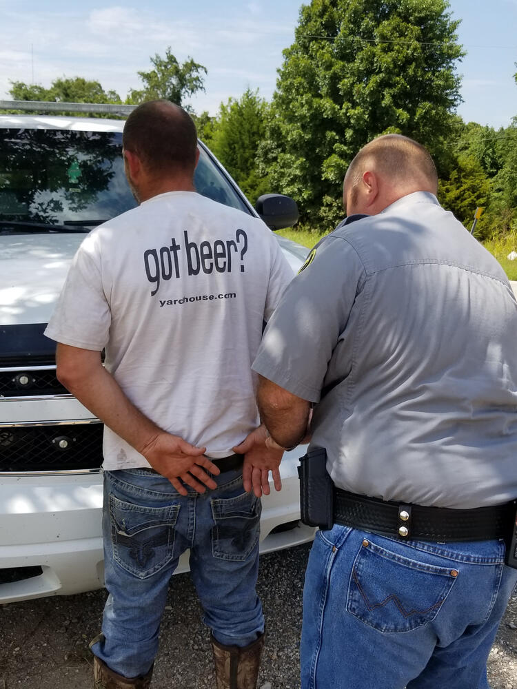 An officer arresting a man. The man is facing the officers vehicle.